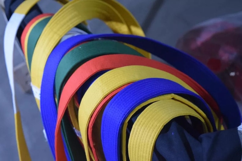 May include: A collection of martial arts belts in various colors, including white, yellow, green, red, blue, and black. The belts are rolled up and stacked on top of each other.