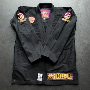 Puede incluir: Gi de Jiu-Jitsu negro con parches bordados en amarillo y morado. El gi presenta el nombre de la marca "SHOYOROLL" en la solapa y la parte inferior delantera. El gi está tendido sobre una superficie gris.
