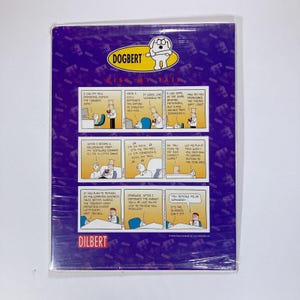 May include: A Dogbert comic strip, titled "Kiss My Tail," is presented in a clear plastic sleeve. The comic features multiple panels with text and illustrations. The background is purple with repeating patterns.