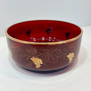 May include: A deep red glass bowl with a gold rim and grape vine design. The bowl has a gold grape vine pattern around the outside. The bowl is likely for serving or display.