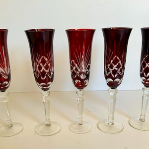 May include: Five ruby red crystal champagne flutes. Each glass has a clear stem and base, with detailed etched patterns on the bowl. The flutes are arranged in a row, highlighting their elegant shape and craftsmanship.