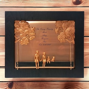 May include: A copper-colored framed artwork with a gold-colored silhouette of a family of four sitting on a swing. The text "The Casey Family" is written above the family, followed by the names "Matt, Amy, Bryant, Gus" and the date "Est. 3.18.2017".