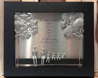 May include: A silver metal framed picture with a silver metal background. The picture shows a family of five sitting on a swing. The swing is made of a black metal bar and rope. The family is made of silver metal and is sitting on the swing. The picture also includes two trees made of silver metal. The text on the picture reads "The Sakhrani Family Amit Manali Shaan Kabir Taj 6th Anniversary Est. January 7, 2018".