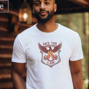 May include: White t-shirt with a graphic featuring a phoenix, flames, and the text "FACE THE FLAME". The design is in shades of red, brown, and orange. The shirt is short-sleeved and has a crew neck.