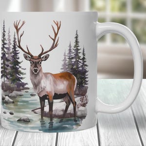 May include: White ceramic mug featuring a watercolor painting of a deer standing in a stream. The deer has large antlers and is surrounded by evergreen trees and rocks. The mug has a curved handle.