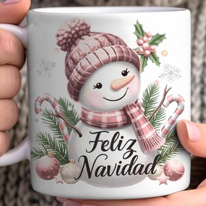 May include: White ceramic mug with a snowman design. The snowman wears a pink and white plaid hat and scarf, and holds candy canes. The mug is decorated with Christmas ornaments, pine needles, and the text "Feliz Navidad".
