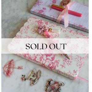 May include: A white notebook with a pink floral pattern and scalloped edges, featuring a pink beaded pen with a bow charm. A matching notepad with a pink ribbon and pen, plus three small floral-themed stickers, are arranged nearby. Text reads SOLD OUT.
