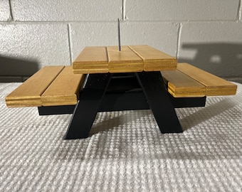 May include: A miniature wooden picnic table with black legs and a brown wood top. The table is made of plywood and has a screw in the center of the top.