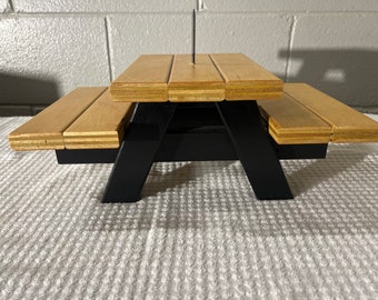 May include: A small picnic table with a light wood top and black legs. The table features a traditional design with a slatted tabletop and attached benches. A single screw is inserted in the centre of the table.