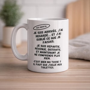 Peut inclure: Mug en céramique blanche avec une anse incurvée. Le mug présente du texte noir en français, dont l'expression "BUG MENTAL". Le texte décrit avec humour un moment d'oubli. Le mug est posé sur une surface en bois.