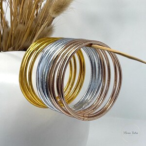 May include: A collection of thin, metallic bangles in gold, silver, and rose gold. The bracelets are stacked together, with a few resting against a white vase with dried wheat stalks. The image is well-lit, highlighting the shiny surfaces.