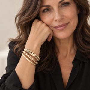 Peut inclure: Un ensemble de trois bracelets dorés portés au poignet. La superposition comprend un bracelet à texture tressée, un bracelet au design torsadé et un bracelet lisse et solide. Chaque pièce présente une finition mate et une couleur dorée uniforme.
