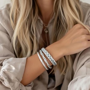Op de afbeelding: Een set van drie zilveren armbanden. Eén heeft een gevlochten ontwerp, de andere twee zijn glad, waarvan één met kleine, ingebedde details. De armbanden worden gedragen op een arm, tegen een neutraal gekleurd overhemd.