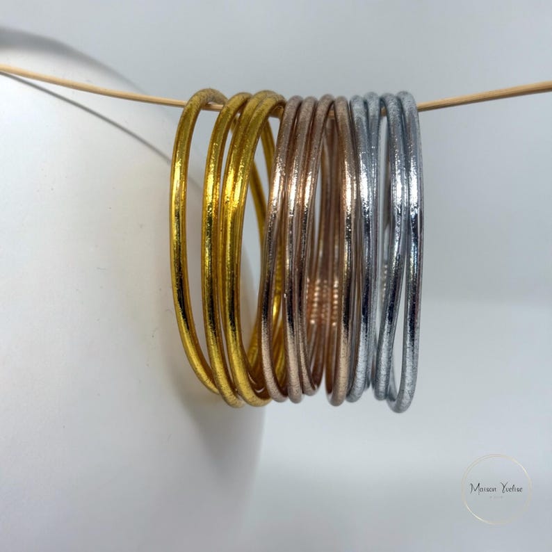 May include: A collection of gold, rose gold, and silver bangles. The bangles are hanging on a thin wooden dowel against a white background. The gold bangles have a shiny finish, while the rose gold and silver bangles have a textured appearance.