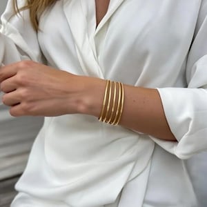 May include: A set of six gold bangles with textured surfaces. The bangles are stacked on a wrist, against a backdrop of a white blouse. The bangles are a warm gold color and appear to be made of metal.