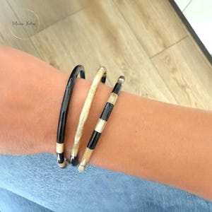 Op de afbeelding: Drie armbanden om een pols. Eén is effen zwart, een andere is lichtbruin en de derde is zwart en wit gestreept. De armbanden zijn gemaakt van een glanzend materiaal. De armbanden zijn op elkaar gestapeld.