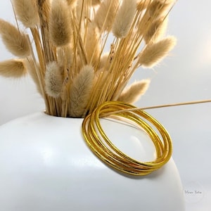 May include: A stack of gold-colored bangles with a textured surface. The bangles are resting on a white surface, with a white vase holding dried flowers in the background.