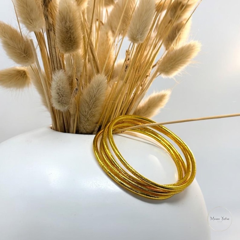 May include: A stack of gold-colored bangles with a textured surface. The bangles are resting on a white surface, with a white vase holding dried flowers in the background.