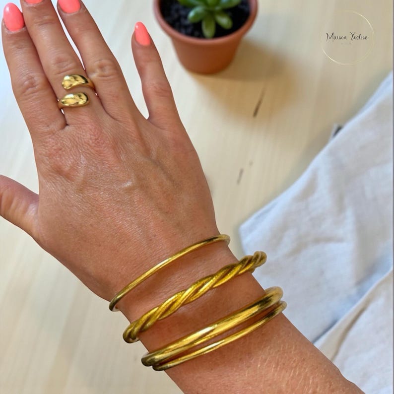 May include: Gold-toned jewelry set featuring a double-band ring and three bracelets. The bracelets include a smooth band, a twisted rope design, and a second smooth band. The jewelry is displayed on a hand with coral-colored nail polish.