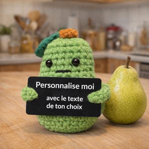 Puede incluir: Una figura de pera de ganchillo verde hecha a mano con un sombrero pequeño, sosteniendo un letrero negro. El letrero dice "Personalízame con el texto de tu elección". Una pera real está al lado.