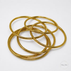 May include: A collection of gold-colored bangles, overlapping and intertwined. The circular bracelets are arranged in a complex pattern, with a shiny, metallic finish. The jewelry is set against a plain white background.