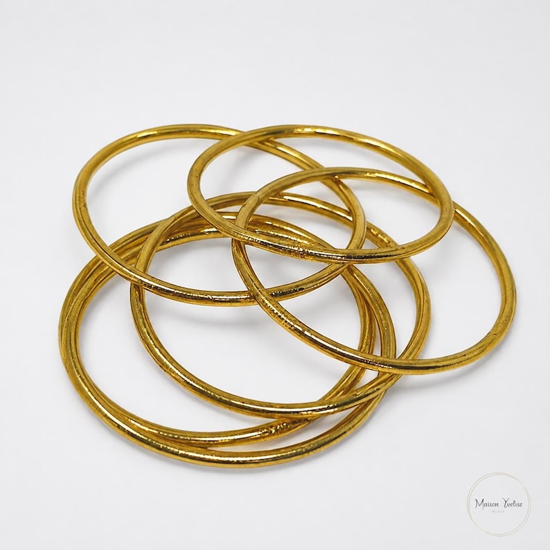 May include: A collection of gold-colored bangles, overlapping and intertwined. The circular bracelets are arranged in a complex pattern, with a shiny, metallic finish. The jewelry is set against a plain white background.