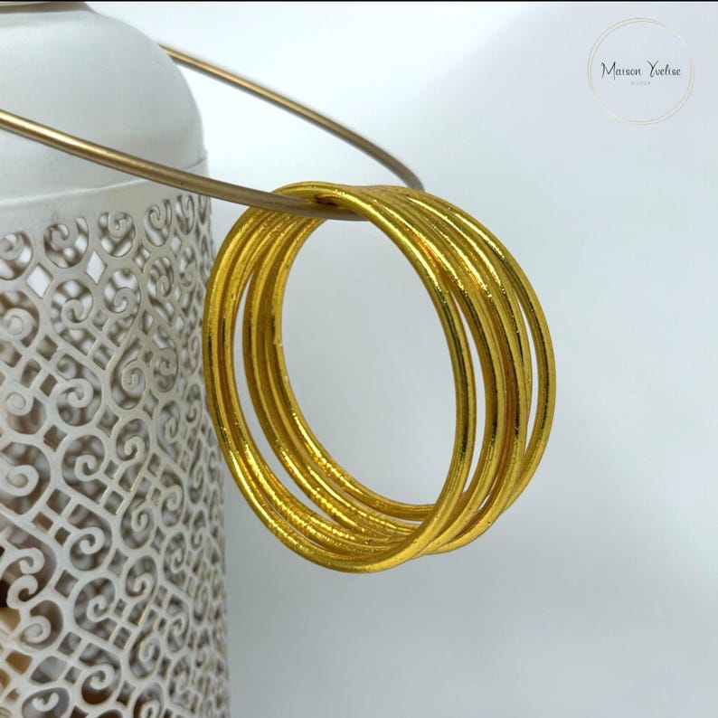May include: A cluster of six gold-colored bangles, each with a smooth, rounded design. The bangles are hanging from a thin, gold-colored metal handle. A white, ornate lantern with a decorative pattern is partially visible in the background.