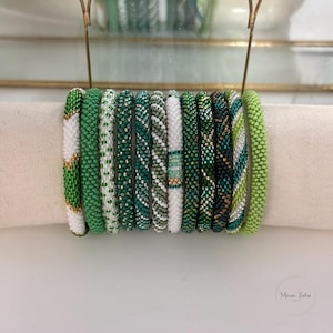 May include: A collection of eleven cylindrical beaded bracelets arranged in a row. The bracelets feature various patterns in shades of green, white, and gold, including solid lime green, striped, and intricate geometric designs made of small glass beads.
