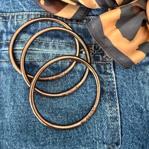 Peut inclure: Trois bracelets joncs circulaires de couleur cuivre avec une finition métallique lisse. Les bracelets sont empilés en se chevauchant légèrement, affichant une forme ronde cohérente et une texture de surface réfléchissante et chaude.