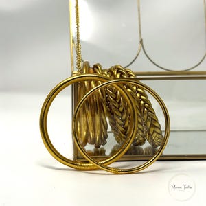 May include: Gold-toned jewelry, including hoop earrings and braided bracelets, are displayed in a glass case. The earrings are large, circular, and smooth. The bracelets have a woven design. The jewelry is set against a white background.