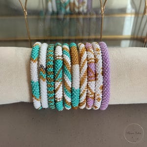 Peut inclure: Une collection de neuf bracelets cylindriques en perles disposés en rangée. Les bracelets présentent des motifs complexes en perles de verre turquoise, blanches, dorées et lavande, avec divers designs géométriques et des bandes unies.
