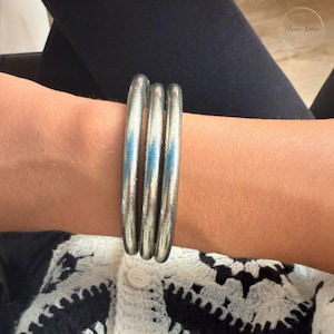 Peut inclure: Trois bracelets argentés sont empilés sur un poignet. Les bracelets ont un design lisse et arrondi et un éclat métallique. L'arrière-plan comprend un haut noir et un cardigan à motifs noir et blanc.