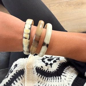 Pode incluir: Três pulseiras artesanais em tons de creme, bege e castanho adornam um pulso. As pulseiras têm um acabamento liso e polido, com padrões e variações de cores únicos. As joias são usadas sobre um suéter com estampa creme e preto.