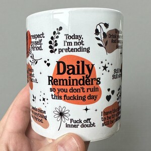 Peut inclure: Mug en céramique blanche avec texte et illustrations noirs et orange. La tasse présente les mots "Daily Reminders" et d'autres phrases comme "Fuck off, inner doubt" et "Today, I'm not pretending."