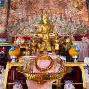 May include: A gold-colored metal tray holds several clear plastic bags containing gold bangles. The tray is held in front of a golden Buddha statue and other religious items. The background features a colorful mural.