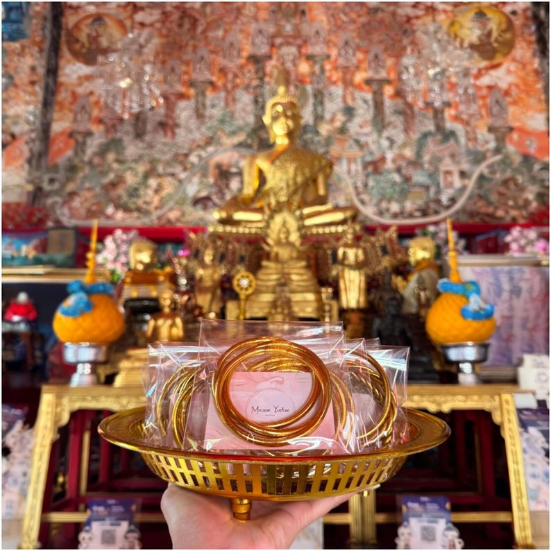May include: A gold-colored metal tray holds several clear plastic bags containing gold bangles. The tray is held in front of a golden Buddha statue and other religious items. The background features a colorful mural.