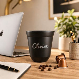 Peut inclure: Une tasse à café noire mate avec le prénom Olivier gravé en écriture blanche. La tasse présente une forme conique et est posée sur une surface en bois, accompagnée de deux capsules de café cuivrées et de quelques grains de café éparpillés.