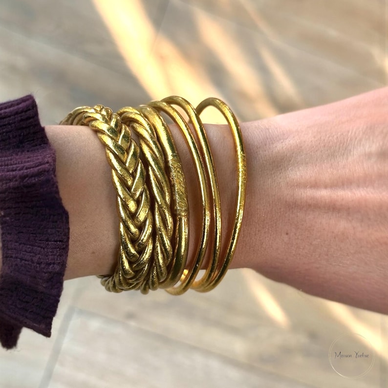 May include: A collection of gold-colored bracelets on a wrist. The bracelets include a braided style and several smooth, round bangles. The wrist is adorned with multiple gold bracelets, creating a layered look. The sleeve of a purple shirt is visible.