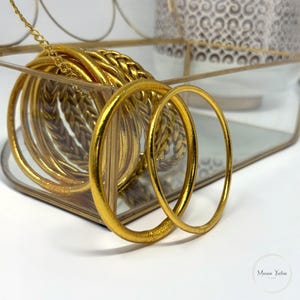 May include: A collection of gold-colored bangles, some with a braided design, are displayed in a clear glass and gold metal tray. The bangles are stacked and arranged, with two bangles in the foreground. A gold chain is attached to the tray.