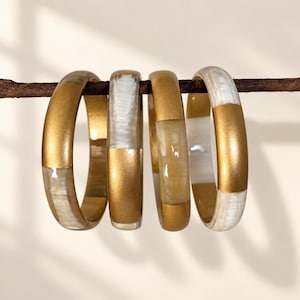 Peut inclure: Quatre bracelets circulaires disposés en rangée, chacun présentant des segments alternés de métal doré mat et d'un matériau blanc translucide lisse avec une texture nacrée et marbrée. Les bracelets sont suspendus à une fine branche brun foncé.
