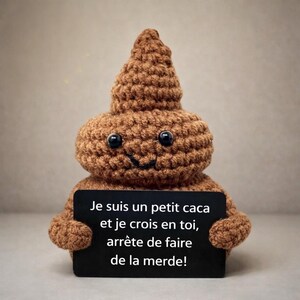 Peut inclure: Un emoji caca marron crocheté avec des yeux noirs en forme de bouton et une bouche souriante. Il tient un panneau noir avec le texte : "Je suis un petit caca et je crois en toi, arrête de faire de la merde!"