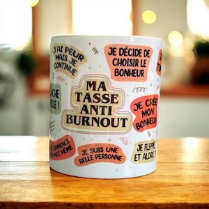 Könnte beinhalten: Weißer Keramikbecher mit verschiedenen französischen Sätzen in verschiedenen Farben. Die Sätze lauten unter anderem "Ma Tasse Anti Burnout", "Je décide de choisir le bonheur" und "Je suis une belle personne". Der Becher steht auf einer Holzoberfläche.