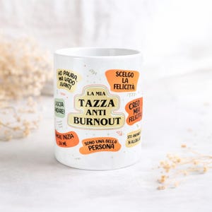 May include: White ceramic mug with the text "LA MIA TAZZA ANTI BURNOUT" in black and orange. The mug has various phrases in Italian, such as "SCELGO LA FELICITÀ" and "SONO UNA BELLA PERSONA", in orange and yellow speech bubbles.