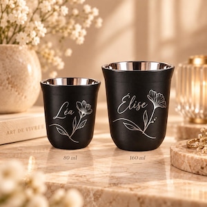 Peut inclure: Deux tasses noires à double paroi avec un intérieur métallique. La plus petite tasse est marquée 80 ml et porte le prénom Léa avec un dessin floral. La plus grande tasse est marquée 160 ml et porte le prénom Élise avec un dessin floral.