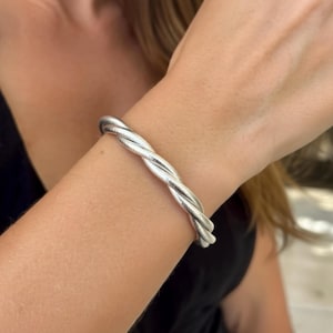 Peut inclure: Un bracelet torsadé argenté. Le bracelet, en métal brillant, est porté au poignet. Le design présente deux brins de métal torsadés, créant un aspect de corde. C'est un bijou simple mais élégant.