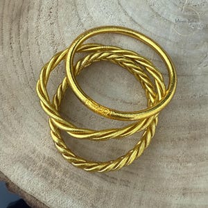 Peut inclure: Trois bracelets dorés avec un motif de corde torsadée. Les bracelets sont empilés, créant un effet superposé. Les bijoux sont en métal brillant. Les bracelets sont de couleur or.