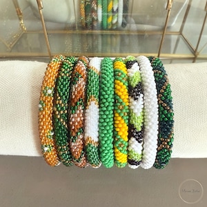 Op de afbeelding: Een collectie van negen cilindrische kralenarmbanden op een rij. De armbanden hebben diverse patronen en kleuren, waaronder oranje, groene, bruine, witte, gele en zwarte glaskralen, elk met een kenmerkende geweven textuur.