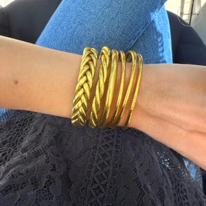 May include: A collection of gold-colored bracelets on a wrist. The bracelets include a thick braided style and several thin, smooth bangles. The jewelry is worn against a background of blue denim jeans and a dark lace top.
