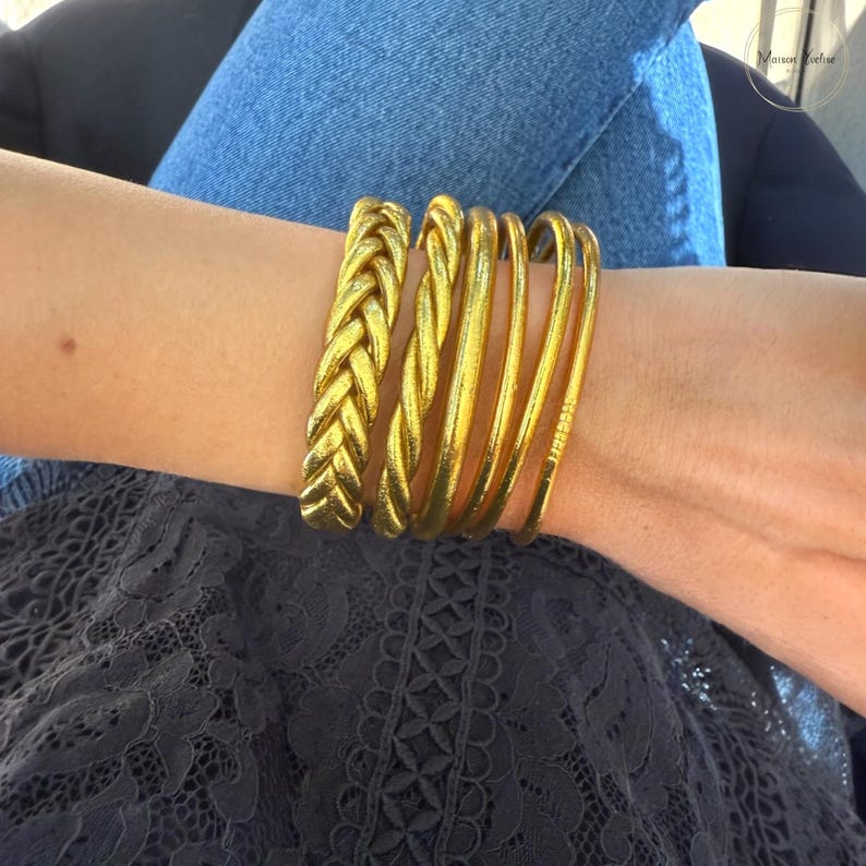 May include: A collection of gold-colored bracelets on a wrist. The bracelets include a thick braided style and several thin, smooth bangles. The jewelry is worn against a background of blue denim jeans and a dark lace top.