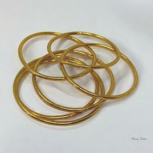 May include: A collection of gold-colored bangles, intertwined and arranged on a white surface. The bangles are circular and appear to be made of metal. The image is well-lit, highlighting the metallic sheen of the jewelry.